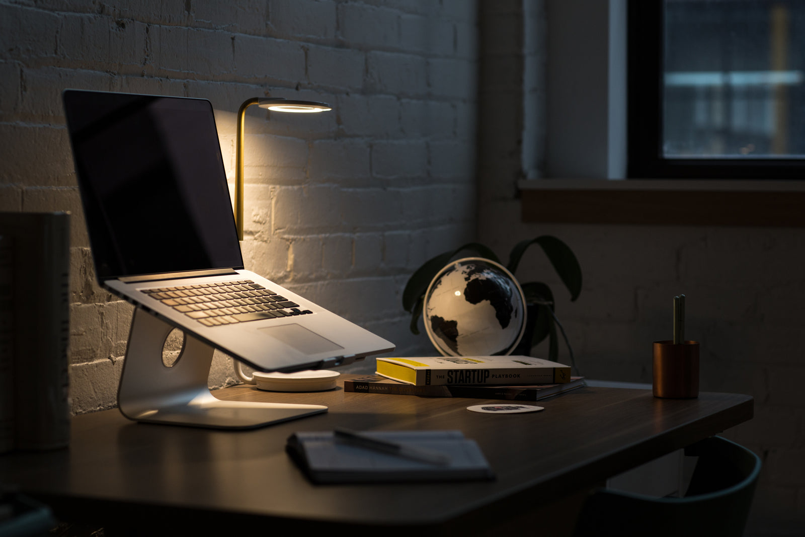 Laptop on a stand on a desk beside a lamp, a globe and books in a dim room